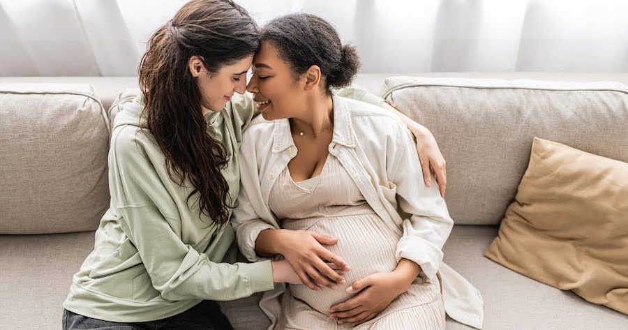 happy pregnant lesbian couple in California, sitting on a couch while holding each other